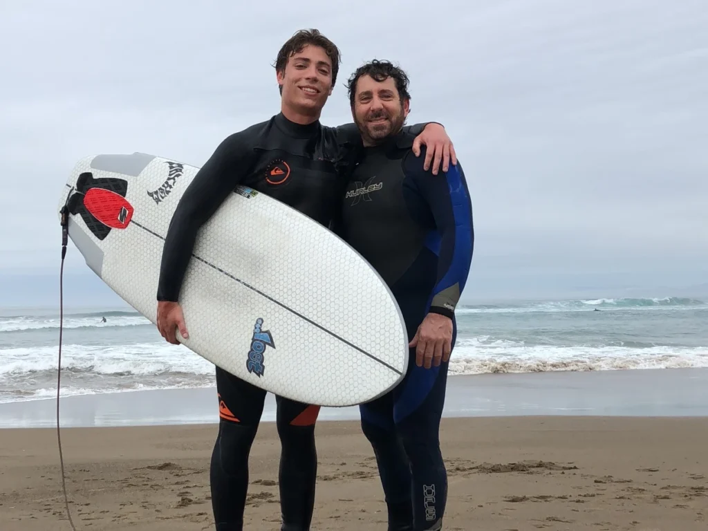 Matthew and father wearing wetsuits, holding surfboard on beach, bonding outside Espo.ai work
